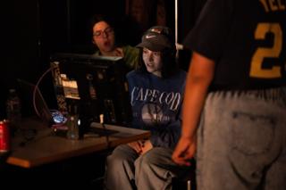Two crew members reacting at the monitor bench, laptops and a mixer spread across the table