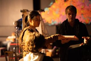Crew members handing off a prop in front of a brightly painted mural, lighting stands behind them