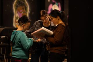 Three crew members leaning together over a script binder, painted murals glowing behind them
