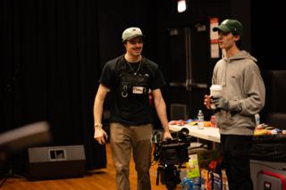 Two crew members walking the camera rig past the craft table, one carrying a coffee
