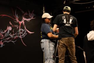 Two crew members working at the camera rig, the pink and red mural spread across the wall beside them