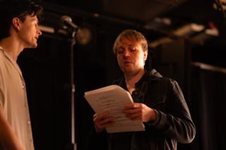 Director reading aloud from a script while the actor listens beside a microphone stand