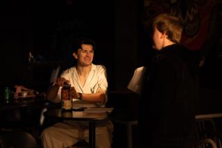 Two actors seated across a small round table in a warmly lit cafe scene