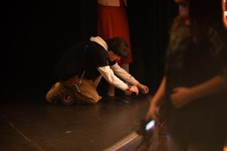 Crew member kneeling to tape down a cable on the stage floor as an actor waits behind