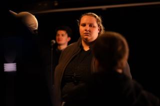 Crew member listening during a discussion on set, an actor at a microphone in the background