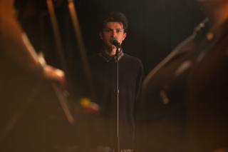 Actor singing into a microphone on stage, framed between silhouetted figures in the foreground