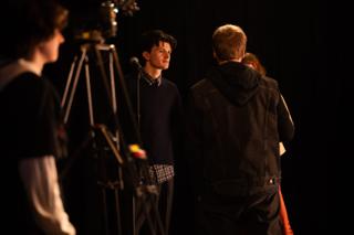 Actor at a microphone on stage while a crew member steps in to give a note, camera rig at the left