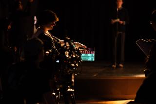 Crew member holding a clapperboard to the lens with the actor waiting on the lit stage behind