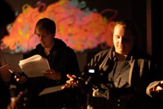 Two crew members reading through script pages in front of a brightly painted mural