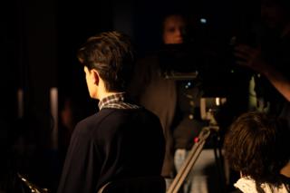 Actor seen from behind facing the camera rig, crew working around them