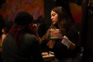 Makeup artist holding a palette while working on a colleague's face beside a painted mural