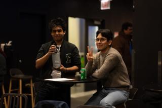 Two crew members at a table during a break, one raising a drink and the other making a peace sign
