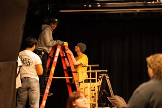 Crew members passing a roll of tape up a stepladder while rigging equipment overhead
