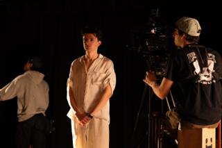 Actor standing on the darkened stage as a camera operator frames the shot