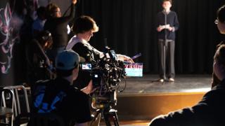 Camera crew at the rig while a clapperboard is held out, the actor waiting on the lit stage beyond