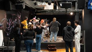 The full set seen from behind the crew, the company gathered around the stage between takes