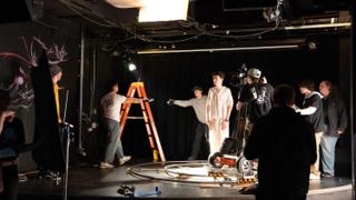 Wide view of the set with the actor mid-scene, lighting stands, a stepladder and dolly track across the floor