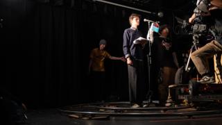 Actor reading from a script at a microphone as the crew work around the dolly track