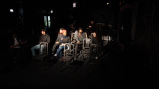 Crew watching from the rows of chairs, stage light spilling across them