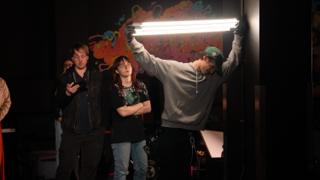 Crew member raising a tube light overhead while two colleagues look on, mural behind