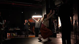 Performer moving across the lit stage in a red skirt, crew watching from the wings