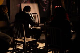 Figure seated alone among rows of chairs in the dark, lit from the side