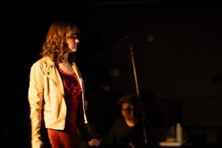 Actor in a cream jacket and red dress standing under a warm stage light